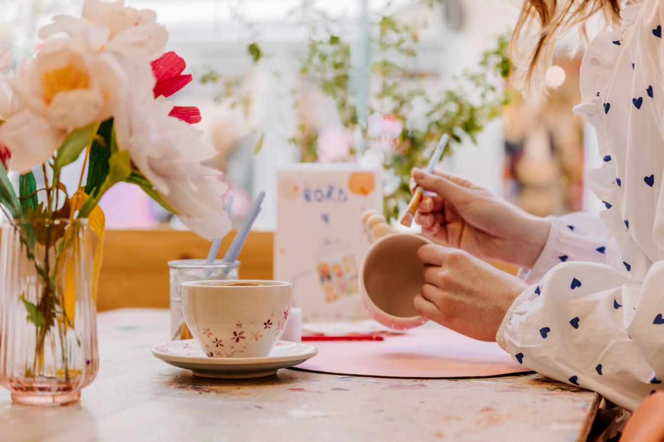 Kvinde maler en kop i kreaværkstedet Kreaværk. Imens nydes en kop kaffe fra cafeen. Butikken er i lyse pastel farver og fyld med blomster og planter