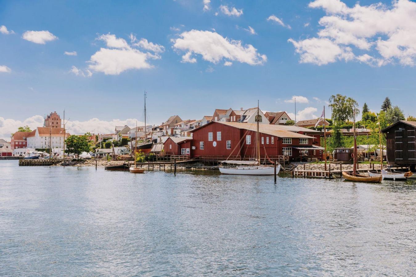 Middelfart Gl. Havn set fra vandet. På billedet ses en gammel havn med træbåde og sejlskibe. Lillebæltværftet, et rødt træhus, ligger ved vandet i havnen. Bagved ses de hængende haver i Middelfart og længere væk kan man spotte Sankt Nikolai Kirke. 