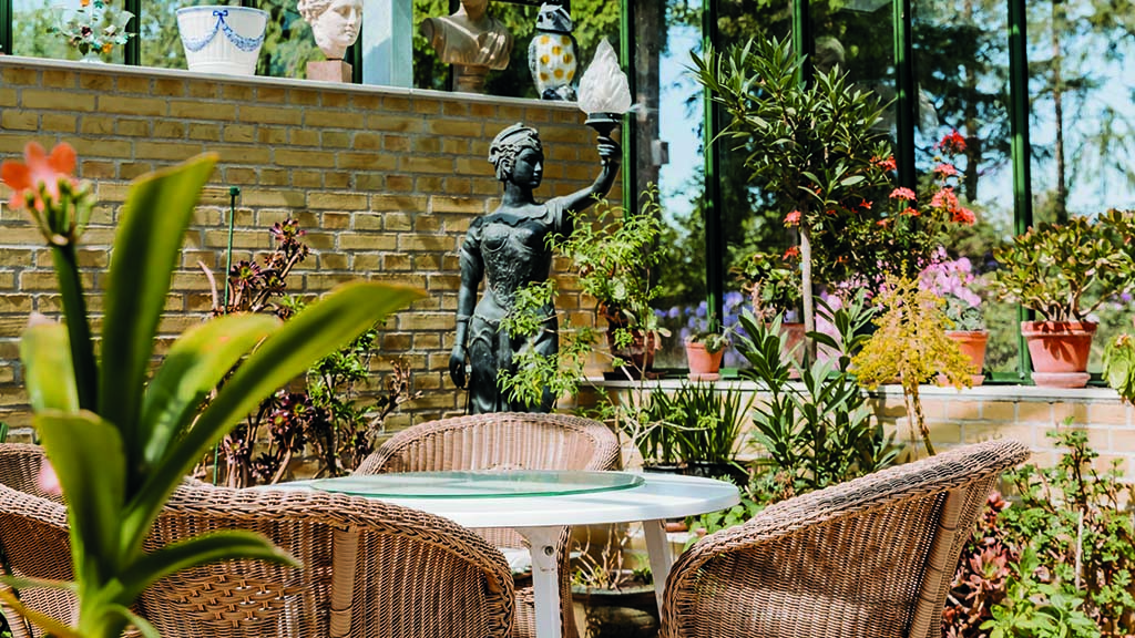 Humlemagasinets cafe i et orangeri fyldt med blomster og statuer. Der er fletstole rundt om et rundt bord. Stilen er Victoriansk. 