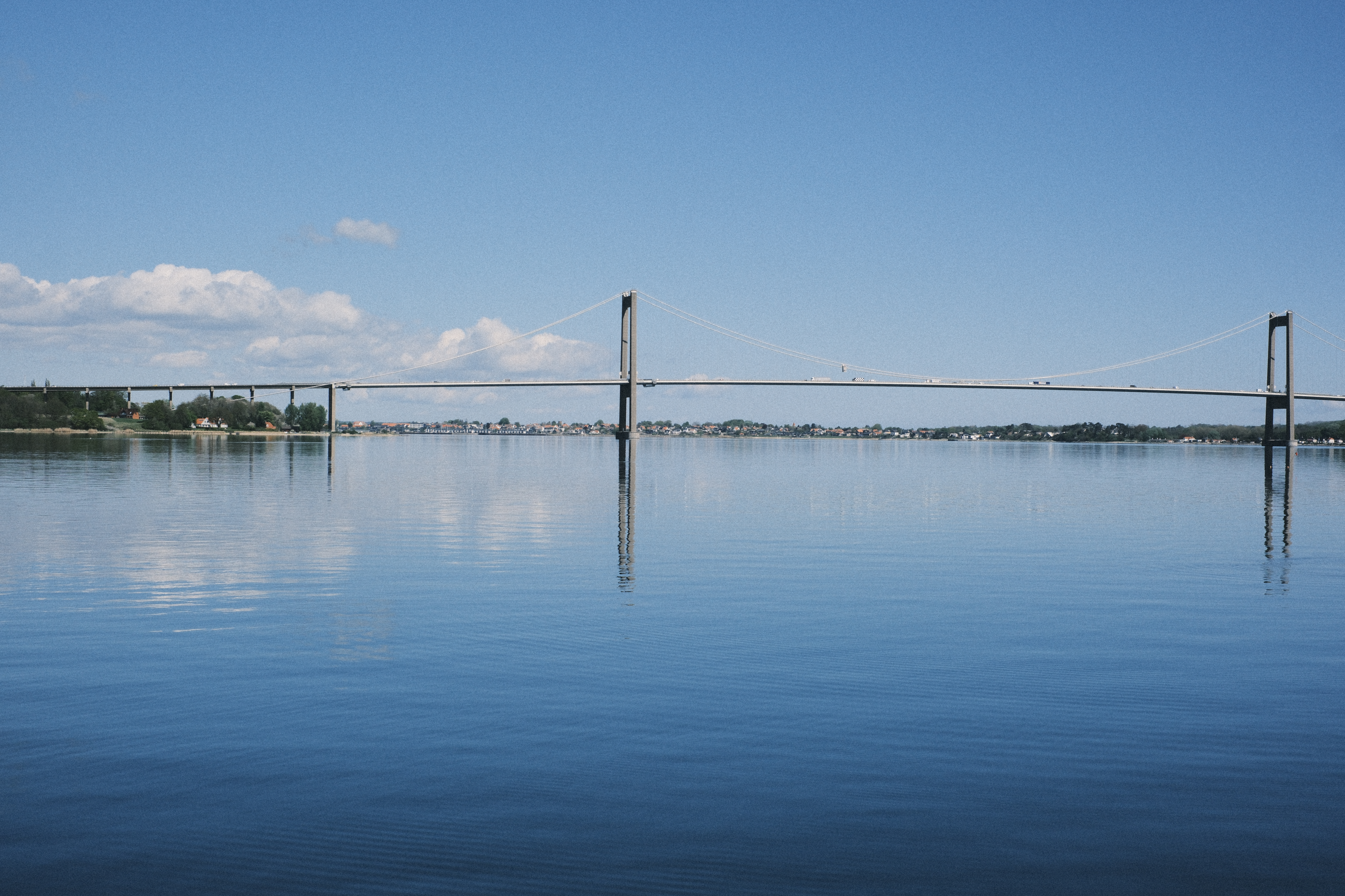 Den Nye Lillebæltsbro set fra havnefronten i Middelfart. Det er en forårsdag og himlen er blå med få skyer. Broen spejler dig i vandet der ligger stille.