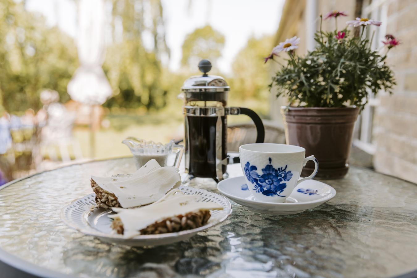 På et cafe glasbord står en tallerken med blondekant. På den er en rugbrødsbolle med ost. Ved siden af er en kop med blåblomst. I baggrunden er en lille Bodum stempelkande. Det er en sommerdag