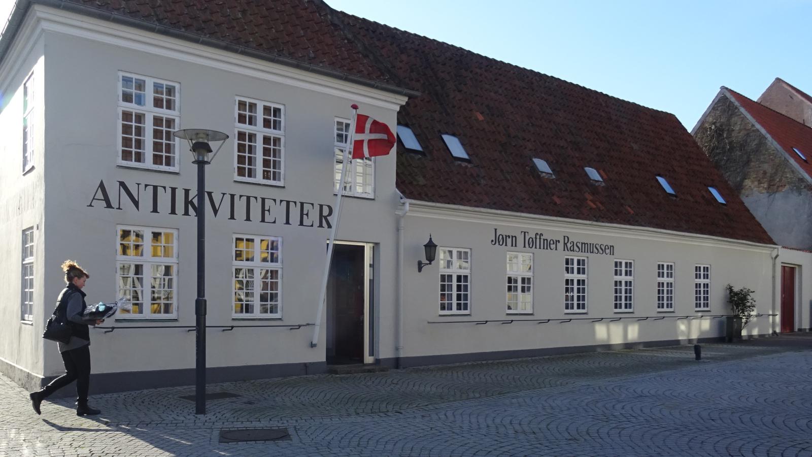 Facaden på Middelfart Antik. En ældre grå bygning med hvide småsprosset vinduer. Ved indgangen hænger et dannebrosflag 