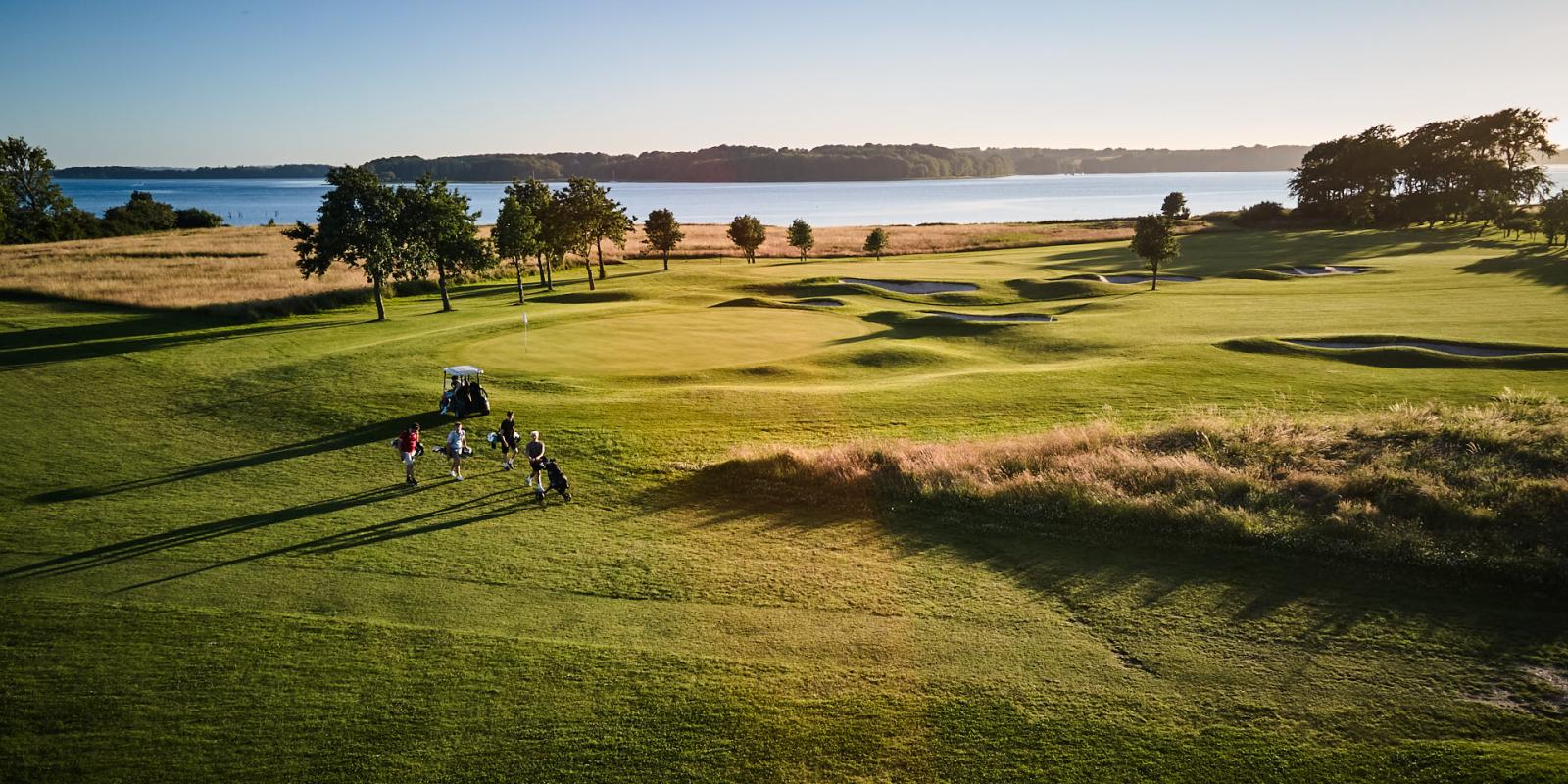 Udsigt over golfbane i Middelfart, Man kan se Lillebælt i baggrunden. Fire golfspillere går hen over banen.