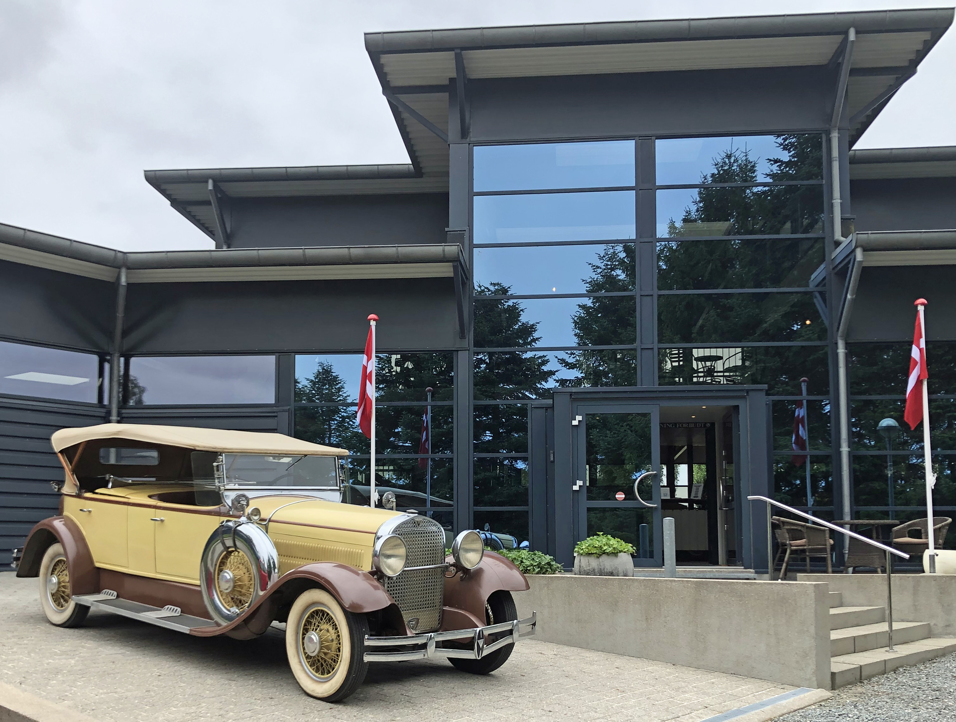En gul veteranbil fra 40erne står foran indgangen til strib automobil museum. Museet indgang har et stort glasparti og to dannebrogsflag ude foran