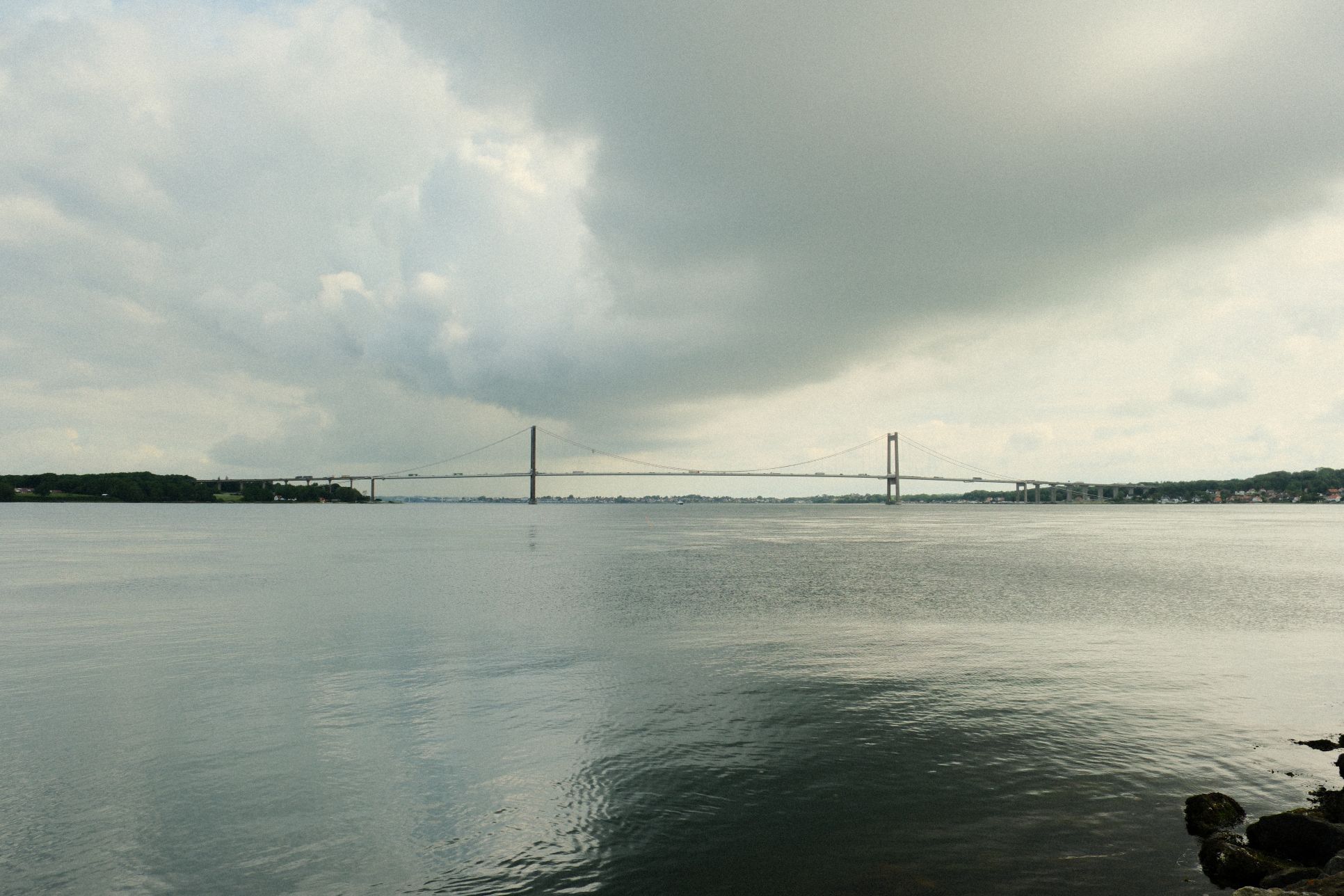 Den Nye Lillebæltsbro set fra havnefronten ved Tråden. Der hænger tunge skyer over broen og vandet ligger stille. 