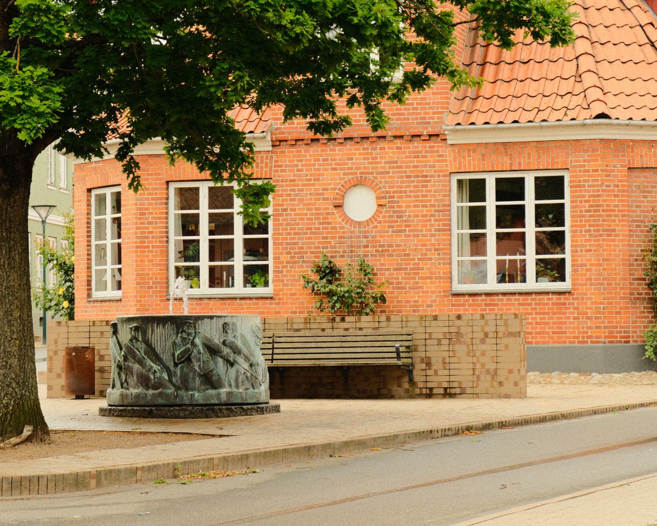 Den gamle bydel i Middelfart. En vej løber langs siden af et hus. regnvand løber i midten af vejen. I venstre side står et gammel træ, bag den er marsvinebrønden og et gammelt rødt murstenshus
