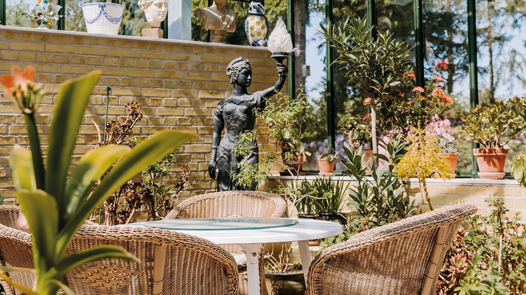 Humlemagasinets cafe i et orangeri fyldt med blomster og statuer. Der er fletstole rundt om et rundt bord. Stilen er Victoriansk.