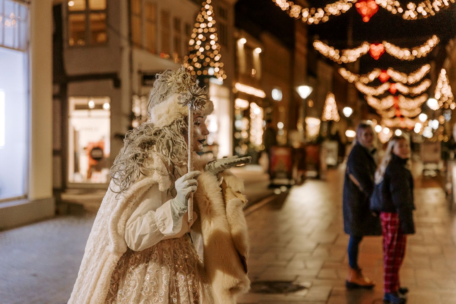 Snedronningen, en dame klædt i hvid kjole, pels, kappe og handsker, laver et luftkys. I den anden hånd har hun en tryllestav. I baggrunden ses Middelfart by klædt i julelys. En mor og datter kigger på snedronningen 
