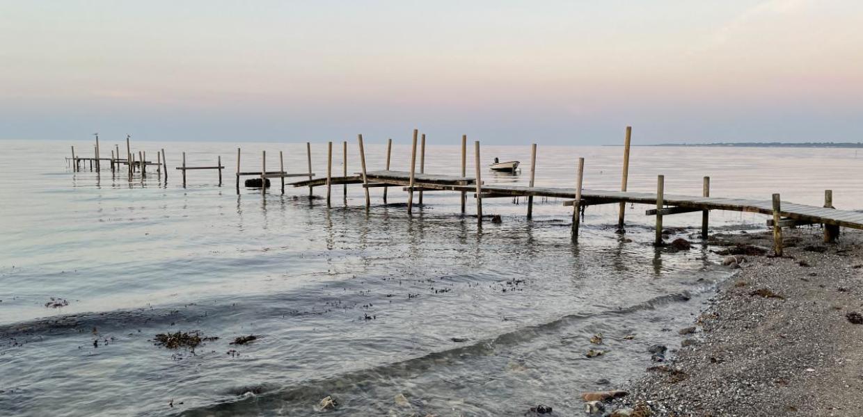 Båd og bro set fra strandkanten med solnedgang i baggrunden